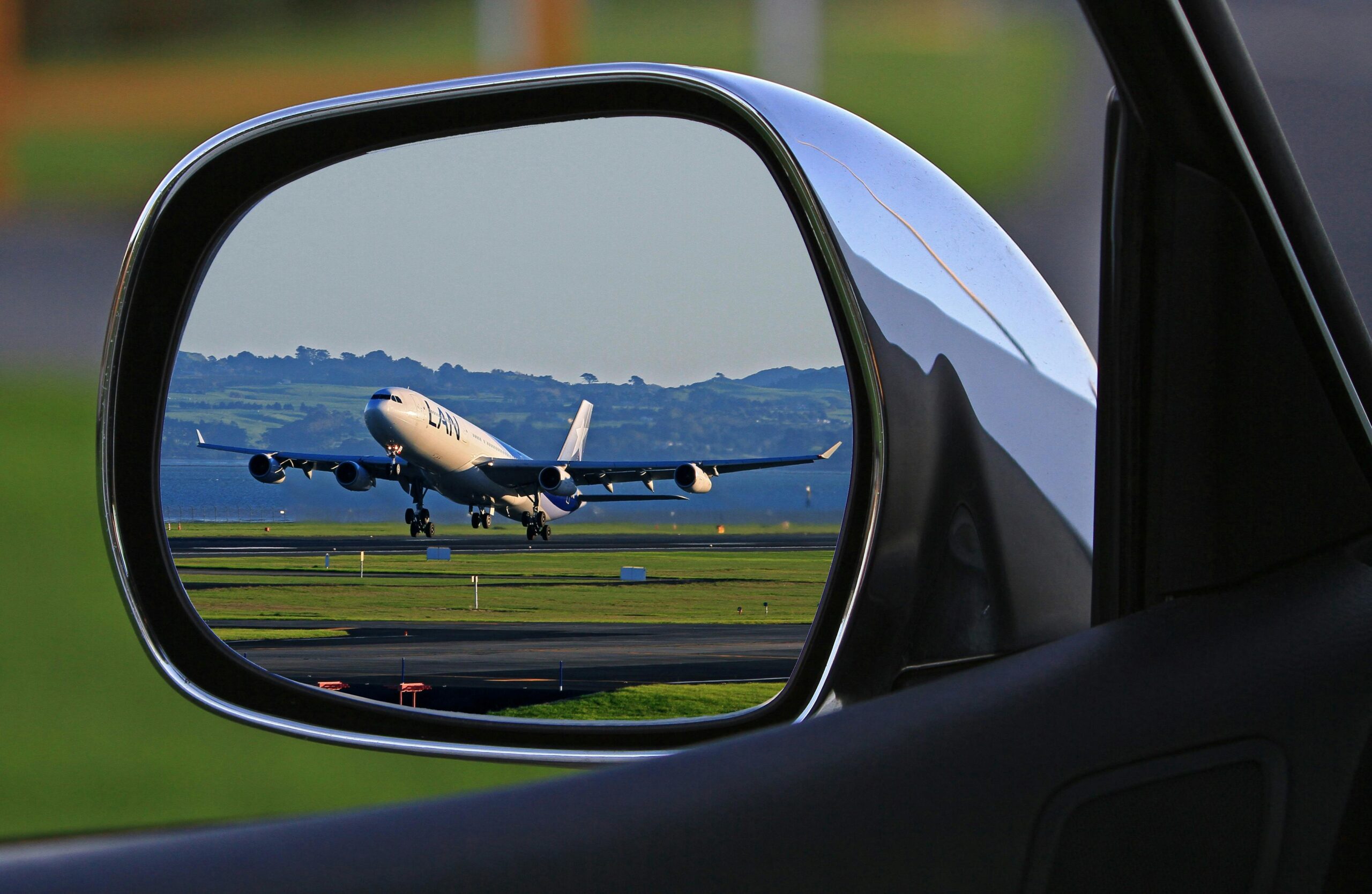A car’s wing mirror reflecting a four-engine airliner taking off from a runway