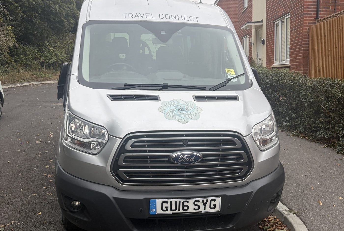 Front of a silver minibus with Travel Connect lettering above the windscreen, parked beside red-brick houses