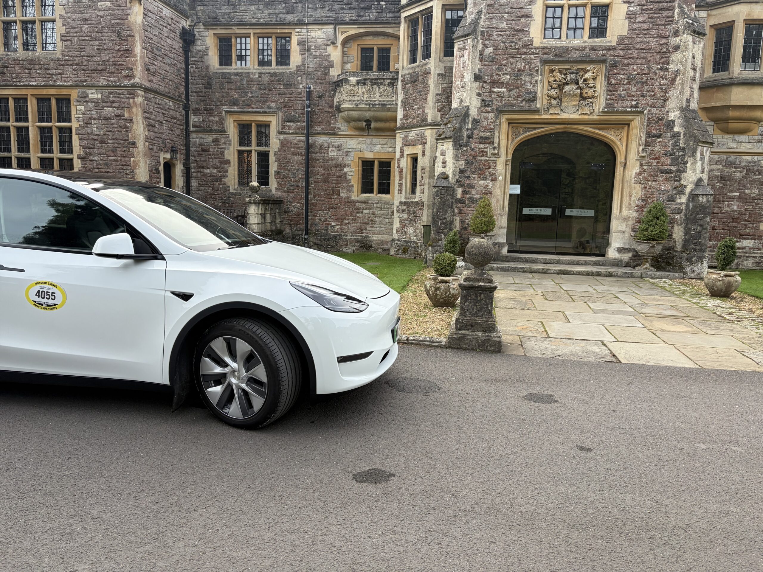 White Tesla Model Y with a private hire licence plate on its door, parked outside the arched stone entrance of a historic manor house