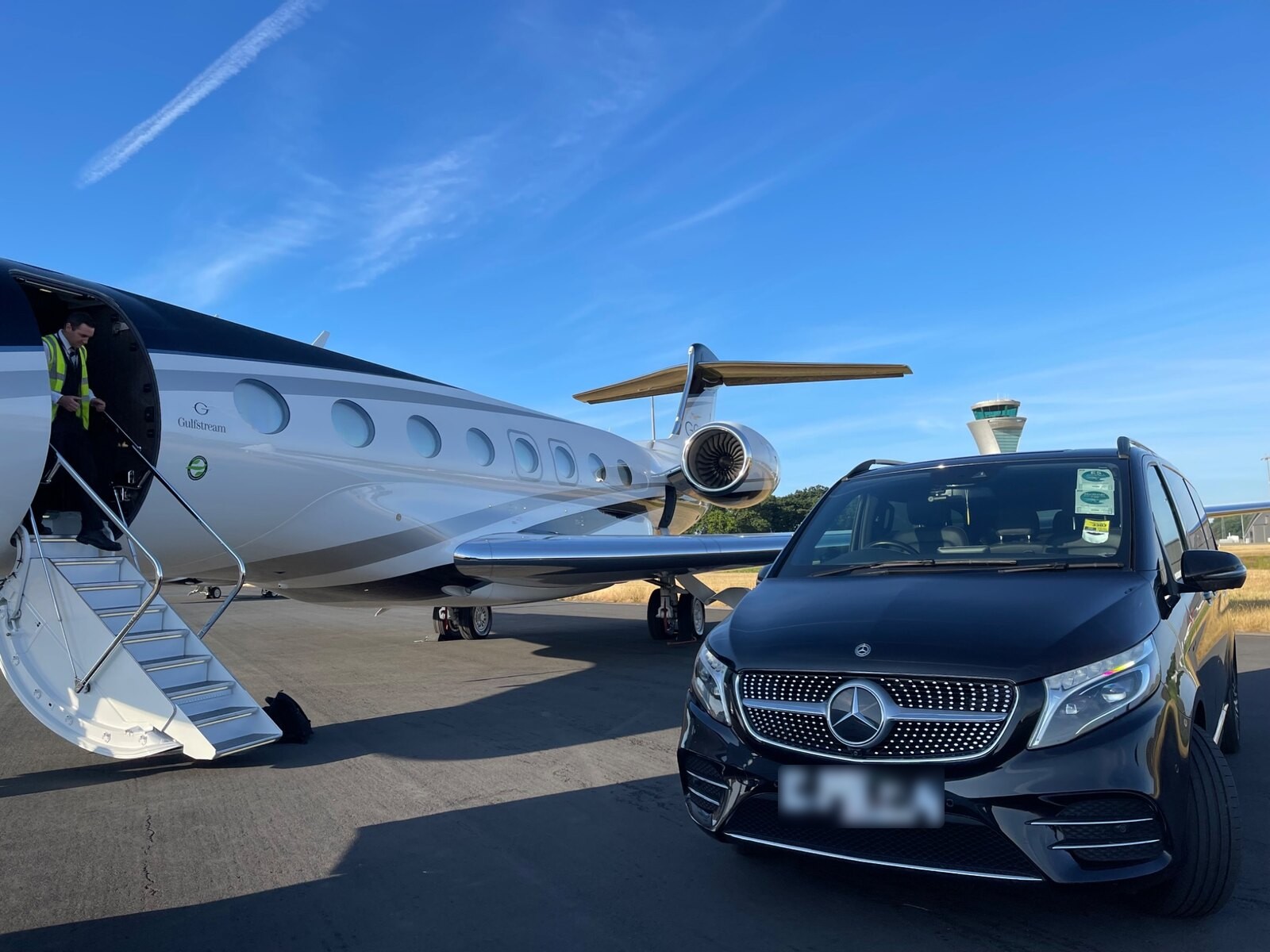 Black Mercedes-Benz V-Class parked beside a Gulfstream private jet on an airfield apron, with a crew member coming down the aircraft steps
